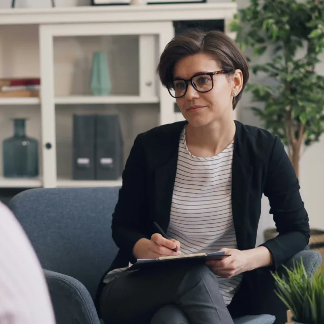 Practitioner seated in session, notebook in hand, facing a client