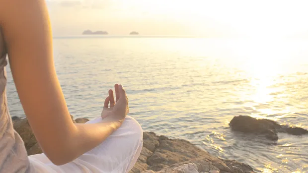 A woman meditates by the sea, facing the setting sun